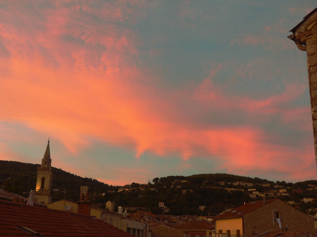 Au loin la vall&eacute;e, le ciel bleu et les nuages orang&eacute;s. Au premier plan, les toits des maisons et sur la gauche le clocher de l'&Eacute;glise Saint Michel de Draguignan.
