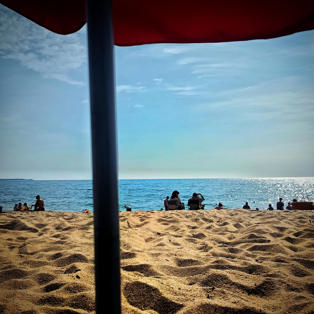 Au premier plan, l&eacute;g&egrave;rement flou le poteau du parasol, juste derri&egrave;re le sable or et la mer bien bleue avec un beau ciel bleu avec de tr&egrave;s l&eacute;gers nuages.